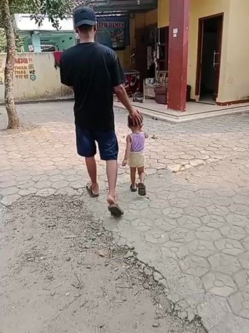 A man in a black t-shirt, blue shorts, a black cap, and flip-flops walks away from the camera, with his right hand resting on the head of a small child. The child, wearing a light purple top and a light yellow skirt, walks slightly ahead of the man, facing away from the camera. They are walking on a paved walkway with hexagonal stones. To the left of the walkway, there is a dirt patch with some leaves and a tree. Behind the man and child, there is a building with a yellow wall and a brown roof. There are various objects visible on and around the building, including signs, a red refrigerator, shelves with items, and some shoes. A mural is visible on the yellow wall to the left. The lighting suggests it is daytime.