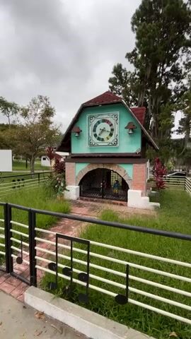 The video shows a small, two-story structure with a red roof and teal-colored upper walls. The upper section features a large, colorful clock face with Roman numerals and moving hands, flanked by two small bells. The lower section has an arched entryway with a black metal gate. Inside the arched area, two small figures, one in blue and one in yellow, are visible. The structure is surrounded by green grass, and a brick pathway leads to the entryway. A black metal fence with musical note cutouts runs along the foreground. In the background, trees, a white building, and a white board are visible under an overcast sky.