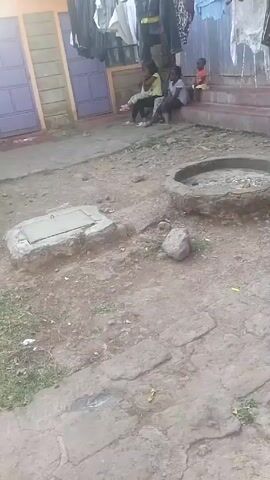 The scene is a residential area with a dirt courtyard. In the background, there's a building with purple doors and clothes hanging to dry above the doorway. Four children are sitting on the steps in front of the building. There's a small concrete slab with a metal handle on it, as well as a circular concrete structure with a hole in the center. The ground is mostly dirt with patches of grass and cracked concrete. A single rock sits in the middle of the dirt area.
