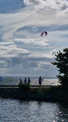 The video shows a body of water with a cloudy sky. There are several people on a walkway or pier, some are standing and some are sitting. There are also several kitesurfers in the water, with kites visible in the sky. The water is choppy and reflects the sky. There is a tree on the right side of the frame. The scene appears to be a recreational area by the water.