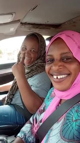 Two women are in a car. The woman on the left is driving while wearing a gray shirt, blue jeans, and a patterned scarf. She makes a peace sign and sticks out her tongue. The woman on the right wears a pink hijab and patterned top. Both women are smiling and wearing seatbelts. Cars can be seen through the windshield in the background.