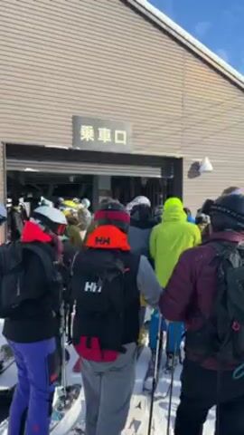 A crowd of people in ski gear are queued up in front of a building. They are all facing the building, waiting in line. Most of them are wearing helmets, ski jackets, and backpacks. They are also holding skis and ski poles. The building has a sign in Japanese above the entrance. The sky is blue and there is snow on the ground. The camera is positioned behind the people in line.