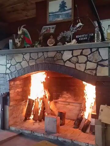 The video shows a large brick fireplace with a roaring fire. There are several logs burning inside the fireplace. Above the fireplace, there is a stone mantelpiece decorated with various items, including a painting of a landscape, a mounted animal head, and some festive ornaments. A sign with text in what appears to be Cyrillic script is also visible on the mantelpiece. The overall environment suggests a rustic or lodge-like setting. The camera pans slightly across the scene.
