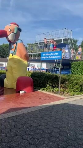 This video captures a lively scene at an outdoor swimming pool in Oelde, Germany. The primary subjects are a group of children and teenagers on a high diving platform, appearing excited and ready to jump. The platform itself is a prominent object, constructed with metal railings and stairs, suggesting it's a designated area for diving or high jumps.

In the foreground, a large, colorful statue of a clown serves as a playful decorative element, adding to the recreational atmosphere. A red helmet rests near the base of the clown, possibly a prop or part of the attraction. The ground around the clown is wet, indicating recent water activity, likely from a splash pad or water feature.

The setting is an outdoor swimming pool area on a sunny day, with a clear blue sky and lush green trees in the background. The presence of other people, including some adults and younger children in the background, suggests a busy public space. The overall mood appears joyful and energetic, characteristic of a summer day at a water park.

A blue sign with white text is visible, displaying the German message: "Die Haftung für Kinder obliegt den Begleitpersonen," which translates to "Liability for children rests with accompanying persons." This sign indicates a safety regulation for the facility. The time of day is likely midday or early afternoon, given the bright sunlight casting shadows.