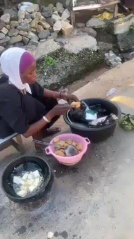 A woman in a black outfit and white hijab is squatting on a small stool outdoors. She is in the middle of cooking, transferring fried dough squares from a pink plastic colander into a black container. A pot on a small stove contains more squares of dough frying in oil.  Behind her is a pile of rocks and what appears to be a drainage ditch.