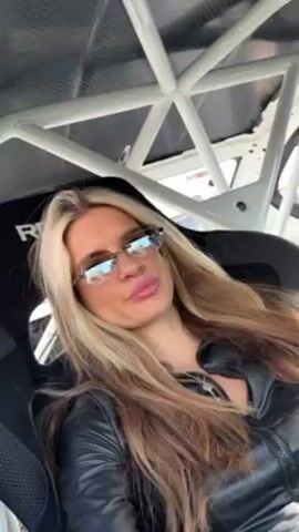 A woman is seated in a Recaro racing seat inside of a vehicle with a white roll cage. She has long blond hair, is wearing tinted sunglasses and a black leather jacket, and is puckering her lips toward the camera. The car appears to be at a racetrack or automotive event, given the sounds of engines and the overall setting. The video is shot from a low angle looking up at the woman.