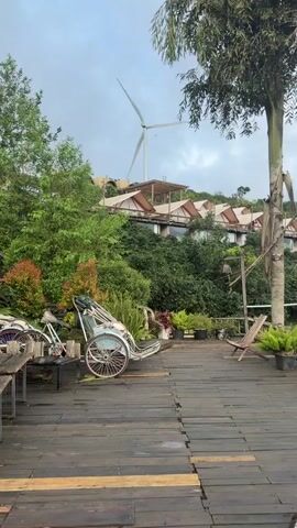 The video shows a static, exterior shot of a wooden deck area with various objects and a scenic backdrop. In the foreground, a weathered wooden deck with planks running horizontally dominates the lower portion of the frame. To the left, there's a small section of a wooden table and a bench. Next to it, a vintage, light blue and white rickshaw is parked. Further to the right, a single wooden lounge chair is positioned on the deck. Beyond the deck, a lush green hillside is covered in trees and shrubs. Several tents with pointed roofs are visible nestled on the slope, suggesting a glamping or resort setting. At the top of the hill, a large white wind turbine with two visible blades stands against a cloudy sky. A tall tree trunk with branches extends down from the upper right corner of the frame. The overall impression is of a tranquil, outdoor location with a blend of natural scenery and resort-style accommodations.