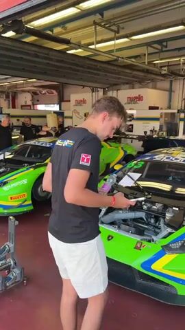 The video shows a pit garage with several racing cars, primarily Lamborghinis, with their hoods open. The cars are green and yellow with racing decals. There are several people in the garage, some of whom appear to be mechanics working on the cars. One person in the foreground is holding a phone and a bottle of water. The environment appears to be a professional racing facility with overhead lighting and garage doors in the background. The overall atmosphere is one of preparation and work on high-performance vehicles.