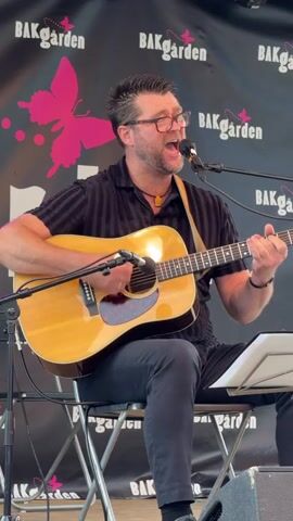 A man with dark hair and glasses is playing an acoustic guitar and singing into a microphone. He is wearing a black striped shirt and dark pants, and is sitting on a folding chair. The background is a black banner with pink butterfly logos and the word "BAKgarden" repeated. The man is strumming the guitar with his right hand and fretting with his left hand. He is looking up and to the right, with his mouth open in song. There is a music stand in front of him. The lighting suggests an outdoor setting, possibly a stage or performance area.