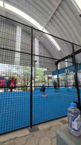 The video shows a paddle tennis match in progress, seen through the wire mesh surrounding the blue court. There are two players on the court, both wearing sportswear. One player is dressed in black shorts and a white top, and the other player is wearing darker clothing. The setting appears to be an outdoor or semi-outdoor covered court, judging by the overhead roof structure. The surrounding environment is visible through the court's fence and shows trees and a glimpse of buildings and vehicles, including a red car. Branding from Columbia is visible on the court support structures. A gray object, possibly a bench or equipment storage, is in the foreground to the right, with a bottle of liquid on it. The camera is positioned outside the court.