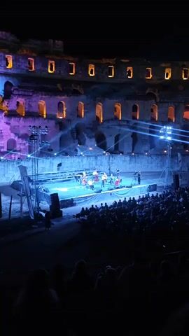 A night concert is taking place in an ancient stone amphitheater. The stage is illuminated with blue and purple lights, and a string orchestra is performing. The audience is seated in the lower tiers of the amphitheater, with their backs to the camera. The upper tiers of the amphitheater are dark, with a few lights visible in the arches. There are stage lights, speakers, and a piano on the stage. The musicians are playing various string instruments, including violins, cellos, and a double bass. One person is playing the piano, and another person is standing and playing an instrument that is not clearly visible. The audience appears to be attentively watching the performance.