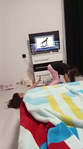 A brightly lit room shows two young children on a bed with a colorful blanket, playing playfully as one child tickles the other. Meanwhile, a girl in pink pants watches a fitness game on a TV sitting atop a white dresser against the wall. Another girl lies on the floor near the bed. A child's potty seat is also visible in the corner. The room features a large window with dark curtains. The children on the bed interact, laughing and moving about on the blanket. The girl watching TV appears focused on the screen, while the girl on the floor remains still.