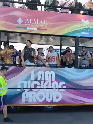 A colorful party bus slowly drives by, full of people celebrating. The bus is decked out with rainbow-themed decorations and logos of ALMAR LGBT Luxury Resort,  Mantamar Beach Club, Aqua Emporio, and Eden. The side of the bus has "I AM FUCKING PROUD" printed in large, bold letters. People are standing and dancing on both levels of the bus, waving their arms and enjoying themselves. A man in a neon yellow vest and blue shorts walks alongside the bus. The sky is overcast, suggesting an outdoor parade or festival.