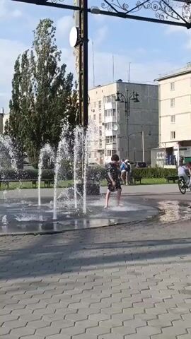 A group of people are walking and riding bicycles past a public fountain. A young person is standing in the fountain, enjoying the water. The fountain is surrounded by a paved area and trees. Buildings are visible in the background under a blue sky.
