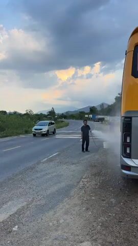 The video shows a man washing a large bus by the side of a road. The bus is silver with a yellow stripe. The man is wearing a black shirt and pants, and he is holding a hose, spraying water onto the side of the bus. A white bucket sits nearby. The road is paved but has a rough shoulder. In the background, there's a car driving on the road, and beyond that, there are mountains under a cloudy sky. The light is soft, suggesting it's either early morning or late afternoon.