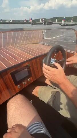 A first-person perspective video shows someone operating a wooden speed boat on a body of water under a partly cloudy sky. The view focuses on the boat's dashboard, which features a digital display showing the speed as 7 km/h and the battery level as 100%. The steering wheel, also wooden, is visible. The boat is moving away from a dock where several flags are planted, and other boats are visible in the distance. The water is calm with some ripples, and the sun is shining. The operator's legs are visible in the lower part of the frame.