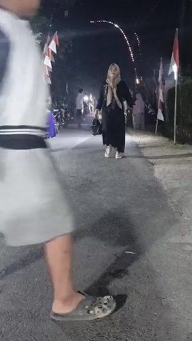 The video shows a street scene at night. In the foreground, a person is partially visible from behind, wearing a grey t-shirt and grey shorts. They are standing on an asphalt road, and their left foot is clad in a dark sandal. Further down the road, a woman wearing a black dress and a beige hijab is walking away from the camera. To her right and behind her, another person in a green headscarf and white pants is also walking. There are flags with red and white stripes on poles lining the street. Decorative lights are strung across the road in the background. Several other people and a motorbike are visible further down the street, and there is a large inflatable structure on the left side of the road. The lighting suggests it is nighttime, and there are shadows cast on the road.