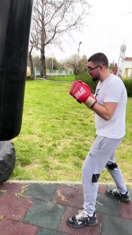 The video shows a young man standing on a play area in front of a black punching bag wearing red boxing gloves. He is wearing glasses, a white Nike t-shirt, and gray sweatpants. He is smiling and sticks his tongue out briefly at the camera. A church and trees can be seen in the background. The ground is covered in green grass and patches of red and green rubber.