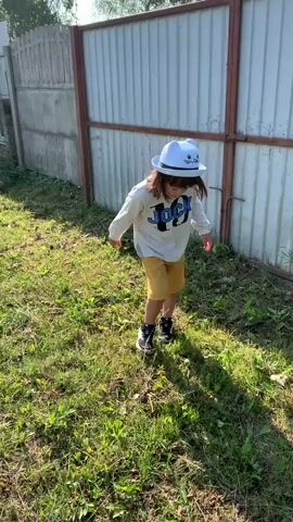 A young child wearing a white long-sleeved shirt with a graphic design and yellow shorts walks across a grassy area. The child also wears a blue hat with a cat design and black and white shoes. The child walks towards a light-colored metal fence with a wooden frame. The sun is shining, creating shadows on the grass.