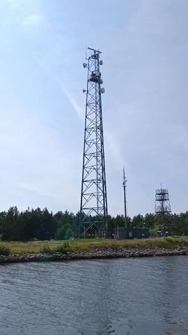 The video shows a telecommunications tower on a body of water. The tower is tall and made of metal lattice. There are multiple antennas and equipment mounted on the tower. In the background, there is a treeline and another, shorter tower. The water in the foreground is choppy. The sky is blue with some wispy clouds.