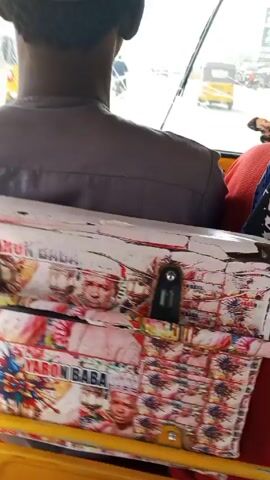 The video shows the interior of a moving vehicle, likely a three-wheeled auto rickshaw, with two passengers visible. The person in the front is wearing a brown top and a patterned cap, while the person in the back is wearing a red outfit with a colorful scarf. A decorated container with images and the phrase "YARON BABA" is placed in the vehicle's interior, with some wear and tear visible. Through the front windshield, other auto rickshaws and pedestrian movement can be observed, suggesting a busy street scene. The windshield appears to be cracked.