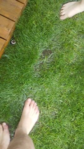 The video shows a top-down view of a patch of green grass, with several bare feet visible. There are puddles of water on the grass, suggesting it might be raining or that the sprinklers are on. A wooden deck runs along the left side of the frame. In the foreground, a person's foot, with blue shorts visible, is stepping into a puddle. Further back, two other bare feet are also on the grass, one to the left and one in the center, closer to the puddles. The scene appears to be outdoors, possibly a backyard or garden, in daylight. The overall impression is of a casual, possibly playful, activity involving water and grass. There is no visible text in the media file.