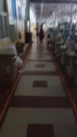 The video shows an interior shot of a factory floor. The camera is moving forward down a wide walkway bordered by tiled squares. On the left side of the walkway, there are some cardboard boxes and what appears to be bags of fabric. Further down on the left wall, there is a fire extinguisher. On the right side of the walkway, there are rows of sewing machines and tables. In the distance, a person in a red coat is walking away from the camera. The lighting is artificial and appears to be overhead fluorescent lights. The overall impression is of an active but not densely populated work area.