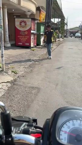 The video shows a person walking on a paved sidewalk next to a street. The person is wearing a dark t-shirt with yellow accents and light-colored pants, and is walking away from the camera. In the background, there are buildings, some with signs, and a few vehicles on the street. The environment appears to be an urban or semi-urban area, likely during the daytime, given the natural light and shadows. The camera is positioned low, suggesting it might be mounted on a vehicle or held close to the ground. There is a portion of what appears to be a dashboard or control panel in the foreground, further supporting the idea that the recording is from a moving vehicle, possibly a scooter or motorcycle. The overall scene is mundane and shows everyday street activity.