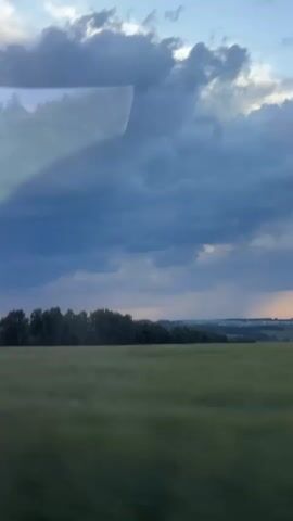 The video shows a view from inside a vehicle, looking out at a landscape. The sky is filled with dramatic, dark clouds, with hints of lighter sky and possibly a sunset visible on the horizon in the distance. In the middle ground, there is a line of dark green trees forming a dense woodland. In the foreground, there is a field of tall, green grass or crops that appears to be moving due to wind or the vehicle's motion. There are reflections on the glass in the foreground, obscuring parts of the view. No people or specific actions are visible, only the natural environment.