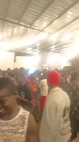 The video depicts a lively indoor gathering of people. Many attendees are dancing and interacting with each other in a large room with a corrugated metal roof and bright overhead lighting. The space is crowded, and various styles of clothing are visible among the participants. Details like a fan and a television screen can be seen in the background, suggesting a social event taking place in a relaxed setting.