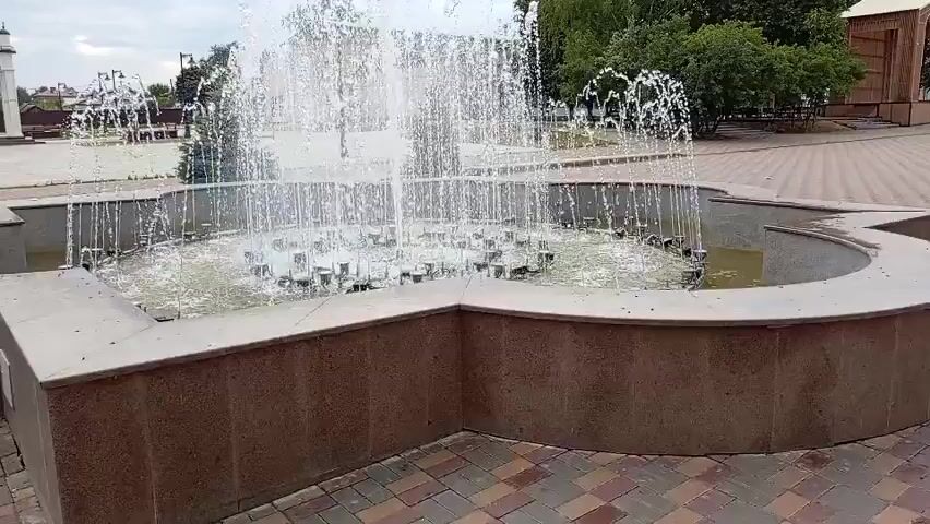 A wide shot shows a fountain in a park on a cloudy day. The fountain has a star-shaped pool with multiple jets of water shooting upwards. In the background, there are trees, buildings, and a paved area.