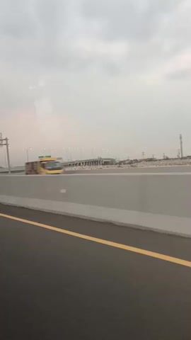 The video shows a view from a moving vehicle, looking out the side window at a highway. The road surface in the foreground is dark asphalt with a yellow line painted on the right side. A gray concrete barrier runs parallel to the road. Beyond the barrier, there are signs, traffic lights, streetlights, and overpasses. In the distance, there are buildings, construction materials, and cell towers. The sky is overcast with gray clouds. A yellow truck and a white car are visible in motion, blurred due to the speed of the vehicle. The overall scene depicts a typical highway environment during cloudy weather.
