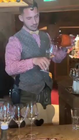 A man in a red-and-white checkered shirt, a vest, and lederhosen is pouring a light orange liquid from a bottle into a wine glass. The man is standing behind a wooden table, on which several empty wine glasses are arranged. There is also a bottle of water, a small bottle labeled "Jägermeister," and other glassware on the table. The background appears to be a rustic restaurant setting with wooden walls and furniture. The lighting is warm and slightly dim.