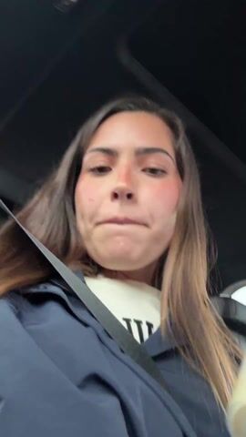 A young woman with long brown hair is seated in a car, wearing a seatbelt and a dark jacket over a white shirt. She is holding a clear blender-style container filled with a light-colored liquid. She takes a sip from the container, then speaks while looking at the camera, which is positioned from a low angle, looking up at her. The car's interior, including the ceiling and parts of the dashboard, are visible. Pop music plays in the background.