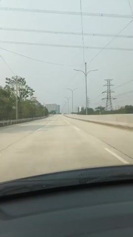 The video shows a view from inside a vehicle, looking forward on a highway. The highway is paved and appears to be empty or has very little traffic. There are multiple lanes in each direction, separated by white lines. On the left side of the highway, there are trees and a guardrail. On the right side, there is a concrete barrier. Several utility poles with overhead wires are visible along the road. In the distance, there are tall buildings and a large electrical pylon. The sky is overcast and hazy, suggesting it may be polluted or foggy. The sun is not directly visible. The timestamp in the video indicates the time is around midnight. The camera is mounted low, likely on the dashboard, capturing the windshield and a portion of the car's interior at the bottom of the frame. There are no people visible in the video. The overall scene depicts an empty highway under a hazy sky during nighttime.