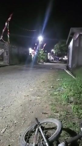 The video begins with a close-up view of a bicycle tire lying on a dirt path at night. The camera then pans up and to the left, revealing a street or alley illuminated by streetlights. Several small Indonesian flags are strung across the path, flapping in the wind. On the right side of the frame, there is a building with a light on inside. The overall scene suggests a residential area at night, possibly decorated for a celebration.