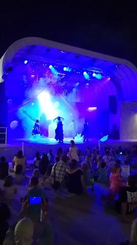 An outdoor stage is illuminated with blue and purple lights. Several performers are on stage, some in elaborate costumes, dancing and moving. There is smoke or fog on the stage, and some large round objects are suspended in the air behind the performers. A crowd of people, including adults and children, are sitting on the ground facing the stage. Some people in the audience are holding up their phones, possibly to record the performance. The overall environment suggests a nighttime event or performance.