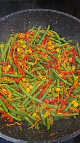 In a close-up overhead shot, a black non-stick pan filled with a colorful stir-fry of green beans, sliced red bell peppers, shredded carrots, and corn kernels is being cooked. Steam rises from the pan, indicating heat. A spatula is used to stir the mixture. Then, a generous amount of cooked, shredded chicken is poured into the pan, adding to the stir-fry. The spatula is then used again to mix the chicken with the vegetables. The camera zooms in slightly as the ingredients are stirred, showing the textures and colors of the dish.