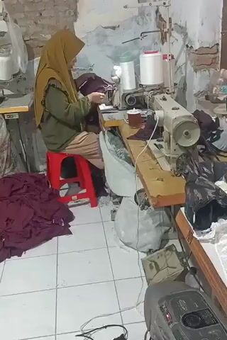 A woman wearing a hijab is sitting on a red stool in front of a sewing machine, operating it. She is dressed in a green long-sleeved top with ruffles on the sleeves and a light brown skirt. To her left, there is a pile of dark red fabric. In front of her, on the sewing machine table, there is a white spool of thread and a white cup with a dark liquid inside. The sewing machine is white and appears to be a standard industrial model. Behind the woman, the wall is made of exposed brick and plaster. To the right of the sewing machine, there are piles of fabric, some of which are black. In the lower right corner of the frame, there is a grey electronic device with buttons and a display, possibly a radio or a music player. The floor is tiled with white tiles. Another sewing machine is visible to the far left of the frame. The overall environment appears to be a workshop or a small production area.