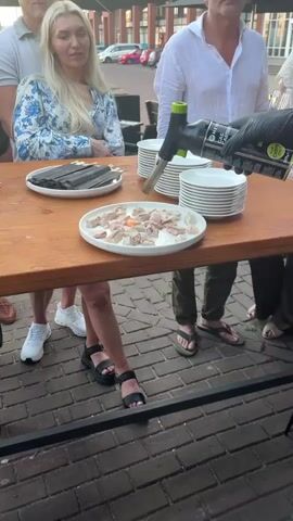 The video captures a close-up view of a table laden with food and plates. In the foreground, a white oval plate holds numerous pieces of raw fish, some of which are being seared with a culinary torch. The torch's flame is visible, creating a dramatic effect as it cooks the fish.

Behind the plate of fish, stacks of white plates are neatly arranged. To the left, another white plate holds what appear to be black chopsticks resting on a black surface. The table itself is made of dark wood.

Several people are partially visible in the frame. A woman with blonde hair and a blue and white patterned top is seated to the left, looking towards the activity. To the right, a person wearing a white shirt and dark pants is standing, holding the culinary torch. Their gloved hand is visible as they operate the torch. The legs and feet of other individuals are also visible, suggesting a social gathering. One person is wearing olive green flip-flops, and another is wearing black sandals.

The setting appears to be outdoors, with a brick-paved area visible on the ground. The lighting suggests it is daytime, possibly late afternoon or early evening, given the warm tones. The overall scene suggests a culinary event or a meal preparation, likely a dinner or a special occasion where guests are being served. The focus on the searing of the fish implies a preparation method that enhances flavor and presentation. The text "HER GAS CARTRIDGE" is visible on the culinary torch.