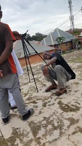 The video shows three men outdoors on a rooftop. One man in an orange shirt and dark pants stands on the left, facing right. In the center, a man in a white robe and dark pants stands behind a tripod with a camera. He is looking towards the right. On the right, a man in a black shirt and gray pants is squatting and looking towards the camera. The background shows buildings with corrugated metal roofs and a tall telecommunications tower. The ground is a worn, light-colored concrete surface. The sky is overcast.