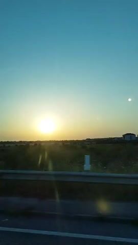 This is a vertical video of a sunset viewed from a moving vehicle. The sun is a bright, white orb low on the horizon, casting golden light across a flat, grassy landscape. A guardrail runs horizontally across the foreground, indicating the perspective of someone in a car or bus on a highway. The sky is a clear, gradient of pale yellow near the horizon transitioning to a bright, pale blue overhead. There are faint lens flares visible. No people are visible in the video. The landscape appears rural with scattered trees and buildings in the distance. The vehicle appears to be moving at a moderate speed.