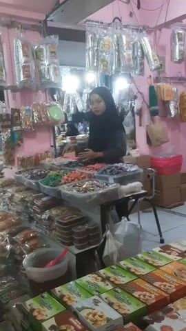 The video shows an indoor market scene with two women. The woman on the right is wearing a black hijab and a dark long-sleeved shirt, standing behind a stall laden with various packaged snacks and candies. She appears to be attending to the stall. The woman on the left is wearing a light brown hijab and a dark long-sleeved shirt, and she is browsing the items on the stall, reaching out to pick up a package. The stall is filled with numerous plastic-wrapped food items arranged on shelves and in containers. The background shows more shelves with packaged goods and fluorescent lighting. The environment appears to be a small shop or a market stall.
