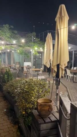 The video shows an outdoor restaurant seating area at night. String lights are draped over trees and pergolas, illuminating tables and chairs. Several large, closed umbrellas are positioned near the seating area. A brick pathway curves through the scene, bordered by planters filled with greenery and decorative pots. A metal railing separates the pathway from a body of water on the left side of the frame. The overall atmosphere is peaceful and inviting.