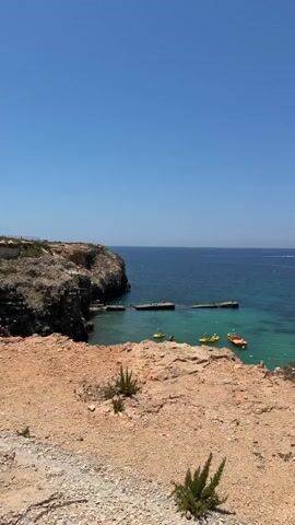 This video shows a scenic coastal view with a small village nestled into a rocky hillside. The water is a clear turquoise, with several small boats and yellow kayaks visible in the bay. The village consists of charming wooden houses with colorful roofs, built on stilts and connected by wooden walkways. The houses are clustered together, some with outdoor seating areas. The landscape is dry and rocky, with sparse vegetation. The sky is a clear, bright blue. There are no discernible people actively engaged in any specific actions within the frame; the video captures a static, picturesque scene.
