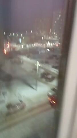 The video shows a view from a high window overlooking a snow-covered street and parking lot in a city at dusk or night. Several cars are parked and some are moving along the road. Streetlights and building lights are visible. The camera view is unsteady and moves slightly, also there are reflections in the window. The window appears to be somewhat foggy or dirty.