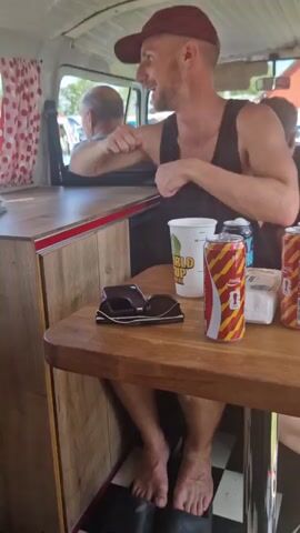 The video shows the interior of a camper van. In the foreground, a man wearing a maroon baseball cap and a dark tank top sits at a wooden table, facing to the left and laughing. He has bare feet. On the table are two cans of soda, a white cup, a pair of sunglasses, and a small white container. Behind the man, another person's head is visible in the background, mostly obscured. The van's interior features red and white polka-dot curtains and wooden cabinetry. Through the window, other vehicles and people are visible outdoors, suggesting an event or gathering, possibly a car show. A Swedish flag is also seen hanging outside the window.
