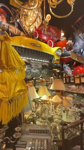 The video pans across a cluttered antique store or flea market. The foreground is dominated by a vintage cash register and an array of silver-plated teapots, pitchers, and other decorative items. A large yellow object, possibly an umbrella or lampshade, is draped over the left side of the frame. Above, suspended from the ceiling, is a yellow classic car, appearing to be a 1955 model based on a visible license plate that reads "34 GASOLIN 1955". The car is decorated with hubcaps. Also hanging from above are various lights, including red dome-shaped lamps, and a large ornate chandelier. On the right wall, a grid of framed pictures, mostly small and uniform, is displayed. Below the car, shelves are filled with an extensive collection of porcelain plates, teacups, and saucers, many with floral patterns. The overall impression is one of abundance and a dense collection of vintage and decorative items.