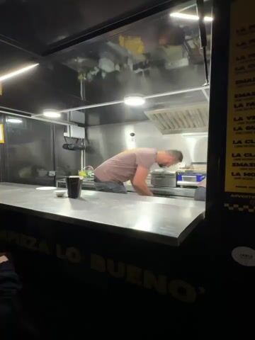 The video shows people lined up outside a food truck at night. The truck is black with yellow lettering that says "EMPIEZA LO BUENO." Inside, a man wearing a brown t-shirt is working at a metal counter. There are containers and a cup on the counter in front of him. A person with dark hair stands to the side, holding a white paper. The camera then pans to show a group of people waiting outside, including a man with curly hair wrapped in a green shawl, who speaks briefly to the camera. The night sky is visible with a faint crescent moon.