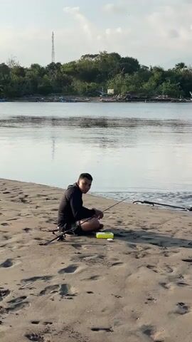 A medium-wide shot shows a man sitting on a sandy beach next to a river, preparing his fishing rod. He is wearing a black hoodie and shorts, with a yellow container beside him. The river has green trees and several boats on the opposite bank. The camera pans right, revealing another man standing further down the beach, looking at his phone. There are clothes and a bag on the sand near him. The sky is bright with some clouds.